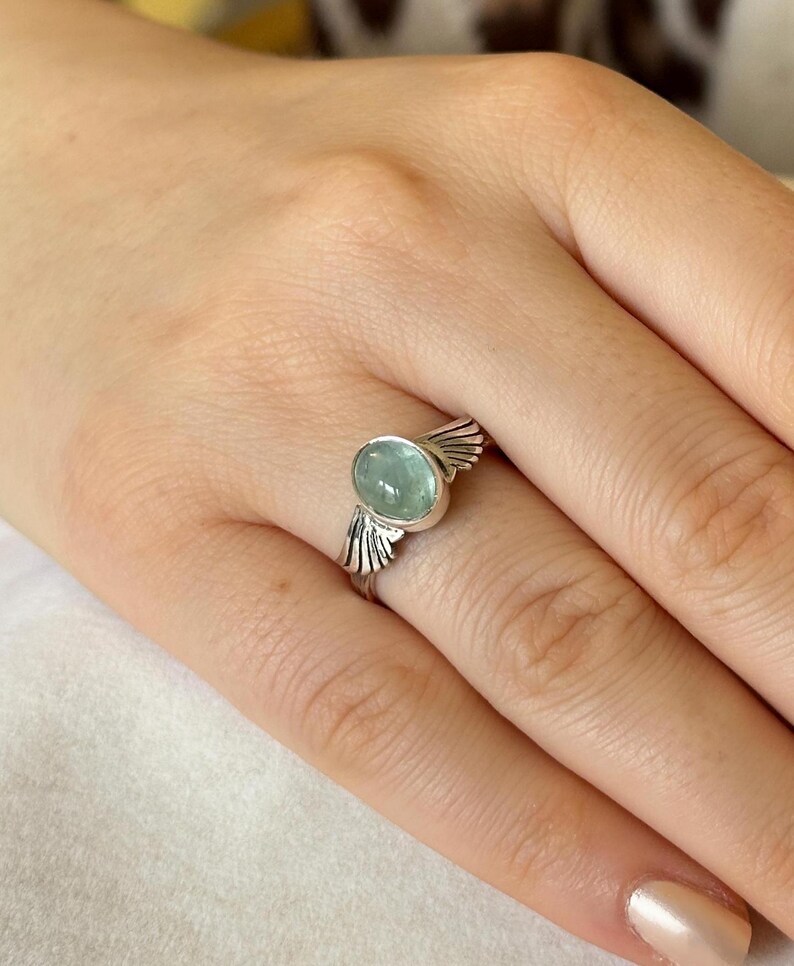 Op de afbeelding: Een zilveren ring met een lichtblauwe ovale edelsteen en vleugelvormige accenten.