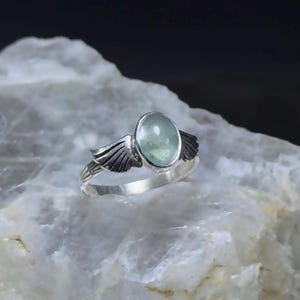 Op de afbeelding: Een zilveren ring met een lichtgroene ovale edelsteen en twee zilveren vleugels aan weerszijden van de steen.