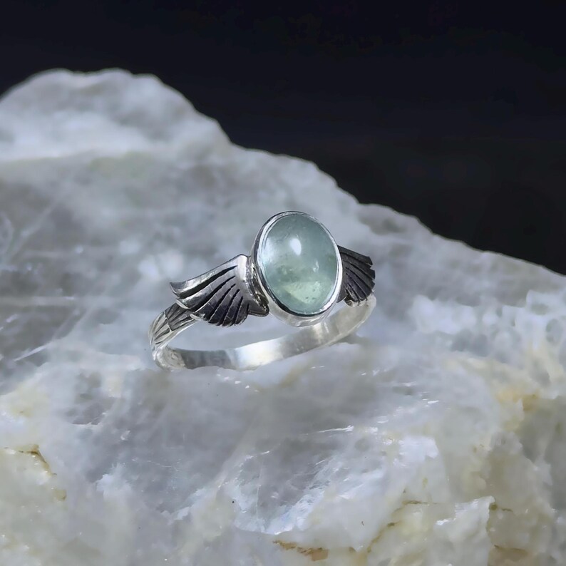 Op de afbeelding: Een zilveren ring met een lichtgroene ovale edelsteen en twee zilveren vleugels aan weerszijden van de steen.