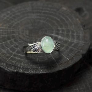 Op de afbeelding: Een zilveren ring met een lichtgroene ovale edelsteen en vleugelvormige accenten.