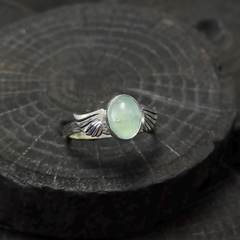 Op de afbeelding: Een zilveren ring met een lichtgroene ovale edelsteen en vleugelvormige accenten.