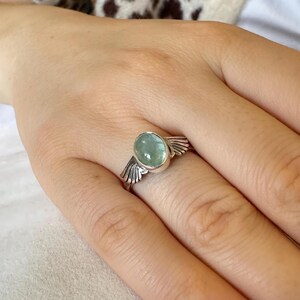 Op de afbeelding: Een zilveren ring met een lichtblauwe ovale edelsteen en vleugelvormige accenten.