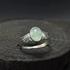 Op de afbeelding: Een zilveren ring met een lichtgroene ovale edelsteen en twee zilveren vleugels aan weerszijden.