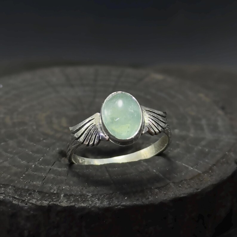 Op de afbeelding: Een zilveren ring met een lichtgroene ovale edelsteen en twee zilveren vleugels aan weerszijden.