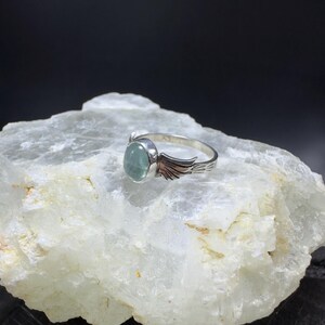 Op de afbeelding: Een zilveren ring met een lichtblauwe edelsteen en zilveren vleugels op de band.