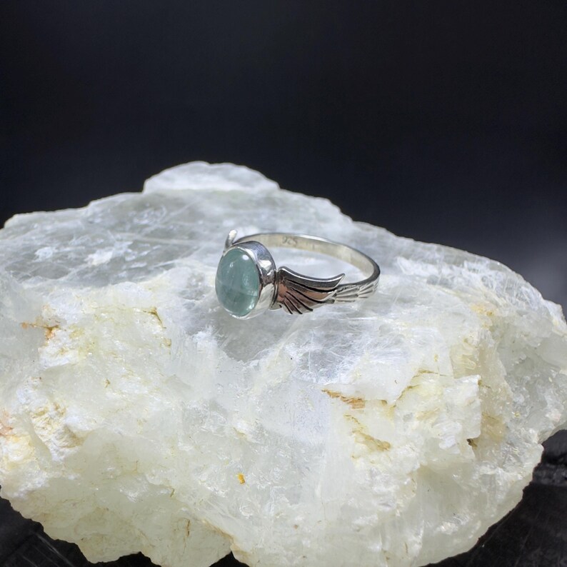 Op de afbeelding: Een zilveren ring met een lichtblauwe edelsteen en zilveren vleugels op de band.