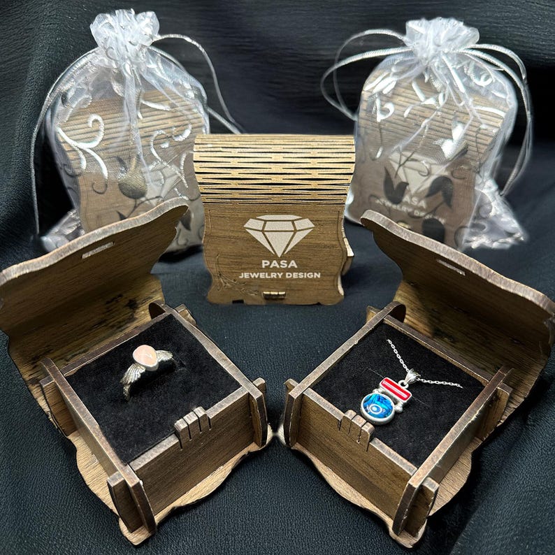 Op de afbeelding: Twee houten dozen met open deksels, die een zilveren ring met een roze steen in de ene doos en een zilveren ketting met een blauwe en rode boze oog hanger in de andere doos laten zien. De dozen zijn gelabeld met "PASA JEWELRY DESIGN".