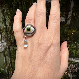 Op de afbeelding: Een zwarte ring met een groot, groen oog en een traanvormig blauw kristal dat eraan hangt. De ring wordt aan een vinger gedragen.