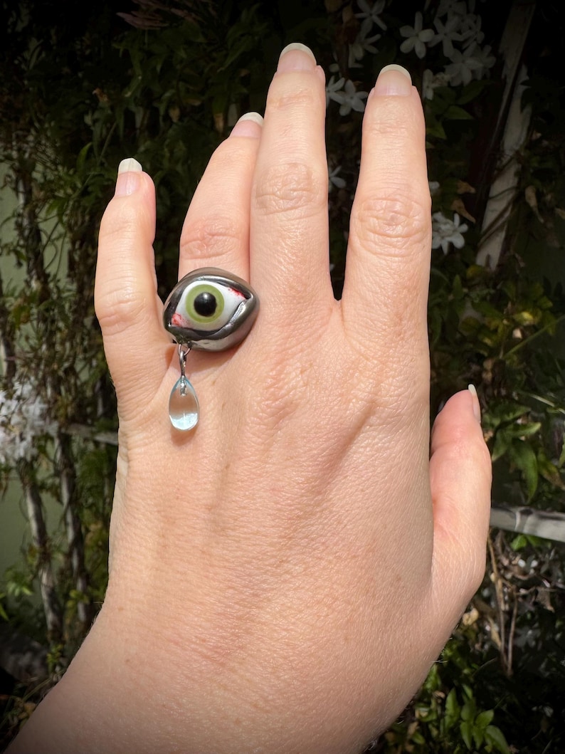 Op de afbeelding: Een zwarte ring met een groot, groen oog en een traanvormig blauw kristal dat eraan hangt. De ring wordt aan een vinger gedragen.