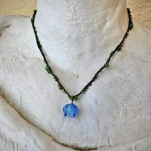 May include: A delicate necklace featuring a blue glass flower pendant. The necklace has a dark brown cord with green beads. The pendant is a light blue, translucent flower shape. The necklace is displayed on a white bust.
