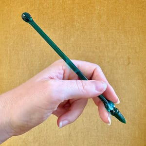 May include: A long, slender, emerald green glass stirring rod with a smooth, rounded bulb at one end and a tapered, pointed tip at the other. The rod is transparent, showing a consistent deep green hue throughout its entire length.