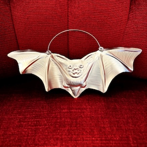May include: A silver-toned metal bat-shaped mold featuring a detailed embossed face, pointed ears, and outstretched wings. A thin, curved metal wire handle is attached to the top of the wings, allowing the object to be hung or suspended.