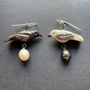 Puede incluir: Un par de pendientes de plata con forma de pájaro con una perla negra y una perla blanca que cuelgan de cada pájaro.