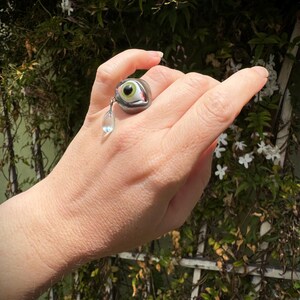 Op de afbeelding: Een zilveren ring met een groot, gedetailleerd glazen oog en een hangende kristallen traan. Het oog is groen met rode aderen en een witte sclera.