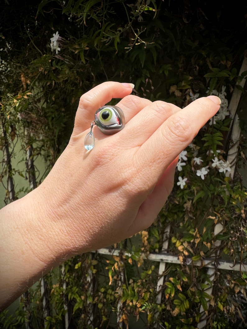 Op de afbeelding: Een zilveren ring met een groot, gedetailleerd glazen oog en een hangende kristallen traan. Het oog is groen met rode aderen en een witte sclera.