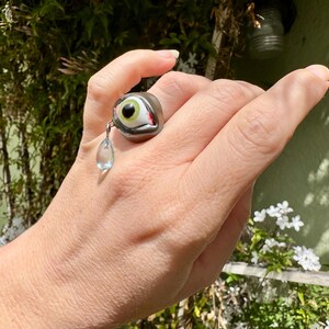 Op de afbeelding: Een zilveren ring met een groen oog en een rode traan die eraan hangt. De ring wordt aan een vinger gedragen.