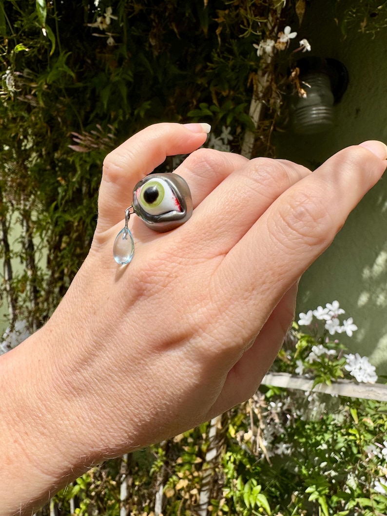 Op de afbeelding: Een zilveren ring met een groen oog en een rode traan die eraan hangt. De ring wordt aan een vinger gedragen.
