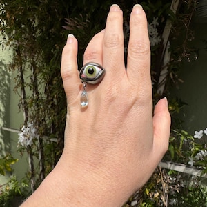 Op de afbeelding: Een zilveren ring met een groot groen oog en een traanvormig kristal dat eronder hangt.