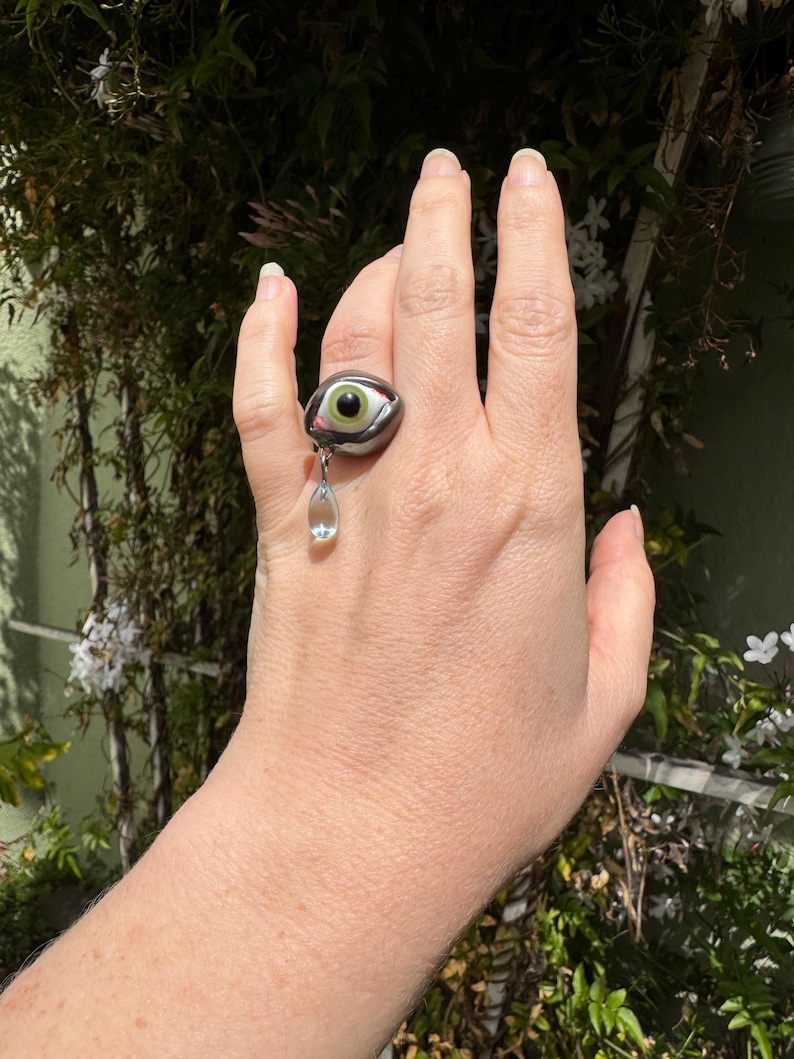 Op de afbeelding: Een zilveren ring met een groot groen oog en een traanvormig kristal dat eronder hangt.