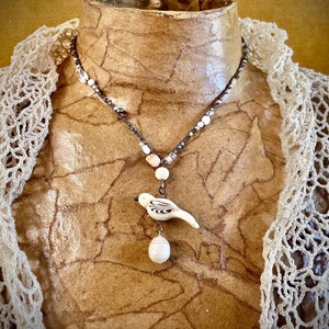 Puede incluir: Un collar con un colgante de pájaro de cerámica blanca y un acento de perla. El collar está hecho con una cadena plateada y cuentas blancas.