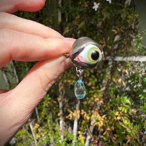 Op de afbeelding: Een zilveren oogbedel met een groene iris en een zwarte pupil, met een rode bloedachtige streep die langs de zijkant loopt. Een traanvormige blauwe kristal hangt aan de onderkant van de bedel.
