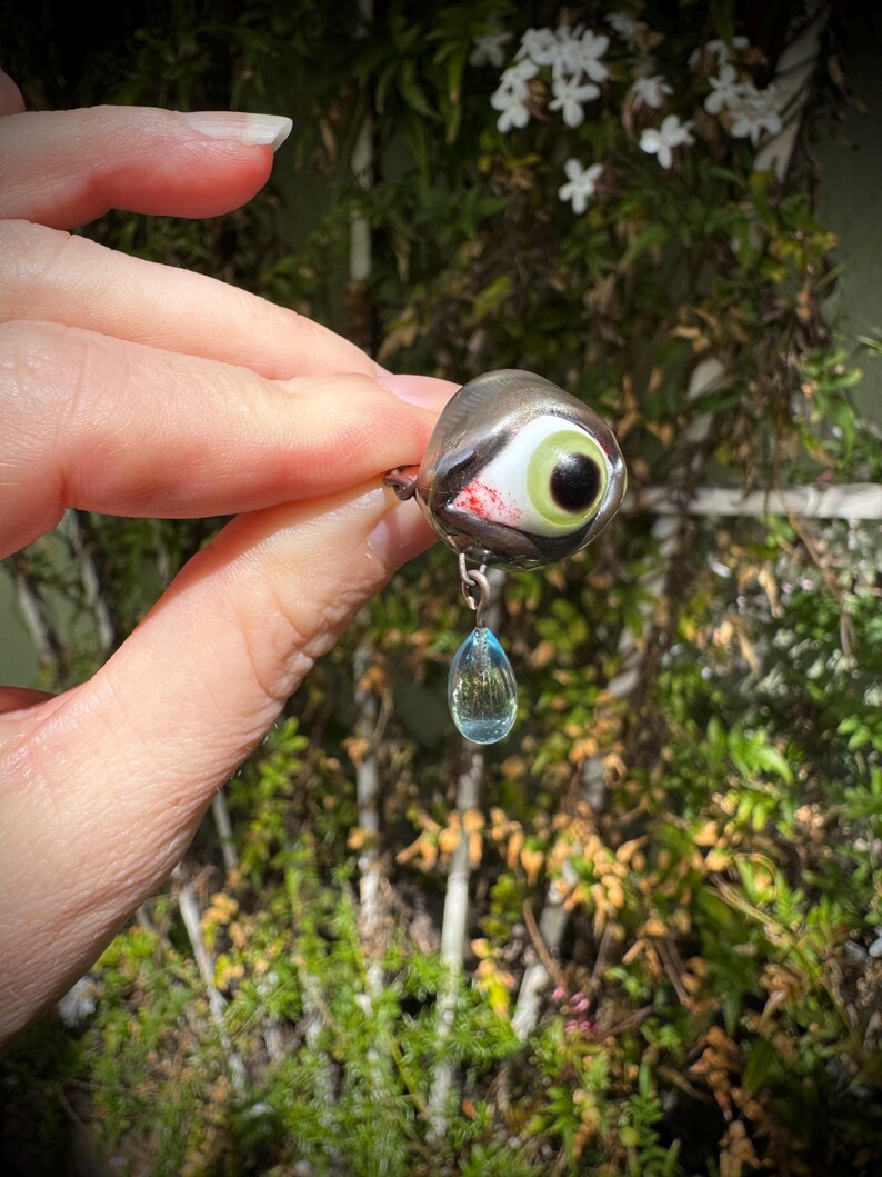 Op de afbeelding: Een zilveren oogbedel met een groene iris en een zwarte pupil, met een rode bloedachtige streep die langs de zijkant loopt. Een traanvormige blauwe kristal hangt aan de onderkant van de bedel.