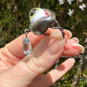 Op de afbeelding: Een zilveren ring met een glazen oog en een traanvormige kristallen hanger. Het oog is groen en wit met een rode ader. De ring is gemaakt van draad en heeft een delicaat ontwerp.