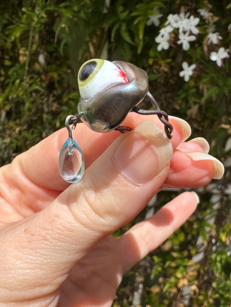 Op de afbeelding: Een zilveren ring met een glazen oog en een traanvormige kristallen hanger. Het oog is groen en wit met een rode ader. De ring is gemaakt van draad en heeft een delicaat ontwerp.