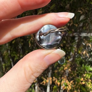 Op de afbeelding: Een zilveren ring met een grote, iriserende, ronde steen die is gezet in een gedraaide draadband. Een kleinere, druppelvormige, heldere steen hangt aan de onderkant van de ring.