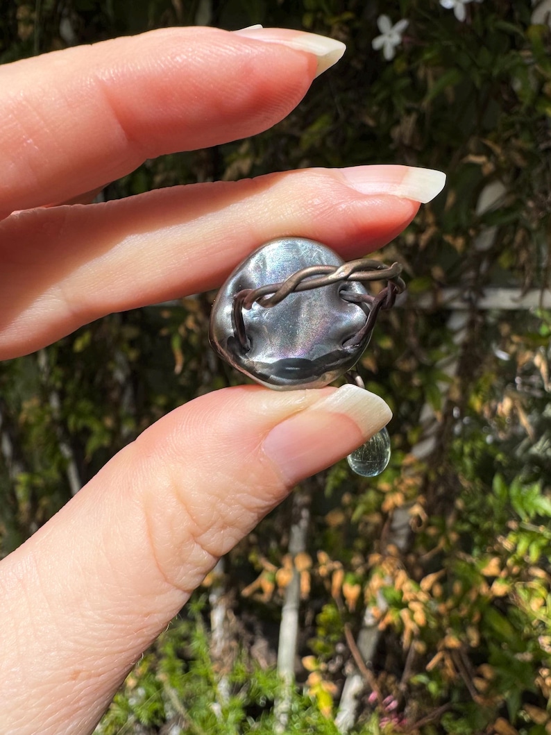 Op de afbeelding: Een zilveren ring met een grote, iriserende, ronde steen die is gezet in een gedraaide draadband. Een kleinere, druppelvormige, heldere steen hangt aan de onderkant van de ring.