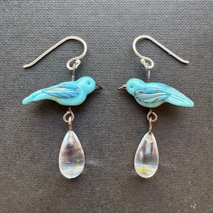 Puede incluir: Un par de pendientes colgantes con abalorios de pájaro de vidrio azul con alas de alambre plateado y gotas de vidrio transparentes.