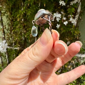 Op de afbeelding: Een zilveren ring met een traanvormige aquamarijn die eraan hangt. De ring is gemaakt van gedraaid draad en heeft een ronde, zilveren bovenkant met een groen-witte steen.