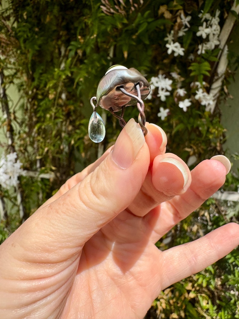 Op de afbeelding: Een zilveren ring met een traanvormige aquamarijn die eraan hangt. De ring is gemaakt van gedraaid draad en heeft een ronde, zilveren bovenkant met een groen-witte steen.