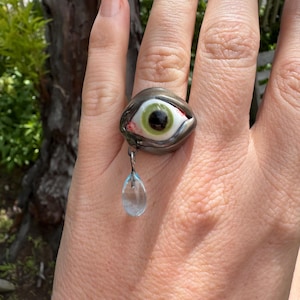 Op de afbeelding: Een zilveren ring met een groen oog en een traanvormige kristallen hanger. Het oog is omgeven door een zilveren zetting en heeft een zwarte pupil. De ring wordt aan een vinger gedragen.