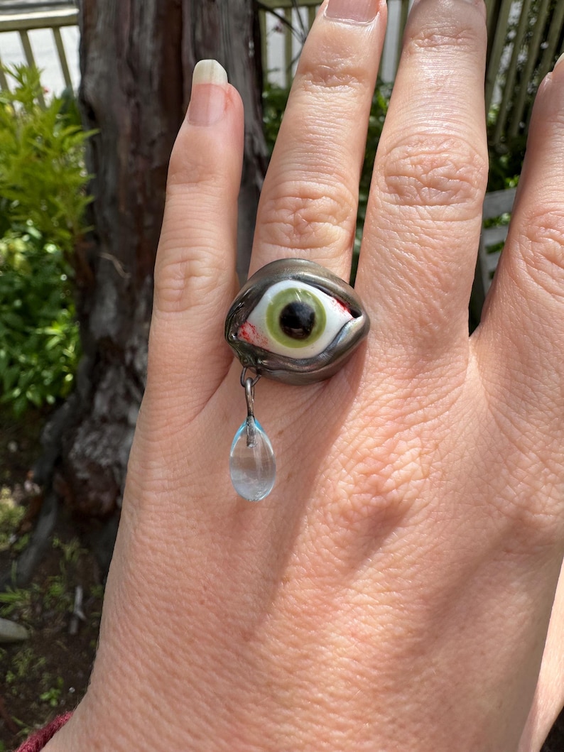 Op de afbeelding: Een zilveren ring met een groen oog en een traanvormige kristallen hanger. Het oog is omgeven door een zilveren zetting en heeft een zwarte pupil. De ring wordt aan een vinger gedragen.