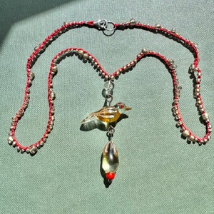 Puede incluir: Un collar de cuentas rojas y plateadas con un colgante de pájaro de vidrio. El pájaro es amarillo con ojos rojos y una lágrima de vidrio transparente colgando debajo.