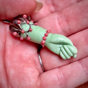 Può includere: Un ciondolo in vetro verde a forma di mano con perline rosa intorno al polso e una montatura in metallo argentato in alto.