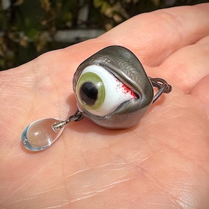 Op de afbeelding: Een glazen oogring met een groene iris en een rode bloedvlek. De ring is bevestigd aan een heldere glazen traanhanger.