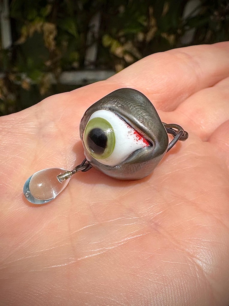Op de afbeelding: Een glazen oogring met een groene iris en een rode bloedvlek. De ring is bevestigd aan een heldere glazen traanhanger.