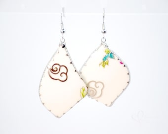Pode incluir: Um par de brincos de porcelana branca com acabamento prateado. Os brincos apresentam um design floral e um padrão de nuvens.