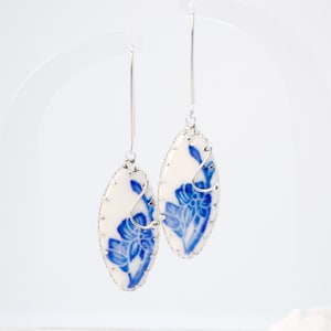 May include: A pair of oval-shaped drop earrings featuring a white ceramic base with a blue floral pattern. Each earring is encased in a silver-toned metal setting with a scalloped edge and hangs from a long, thin silver-toned metal hook wire.