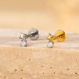 May include: Two star-shaped earrings, one silver and one gold, each featuring a small, clear gemstone. The earrings are presented on a light brown, textured surface. The star design adds a touch of celestial charm.