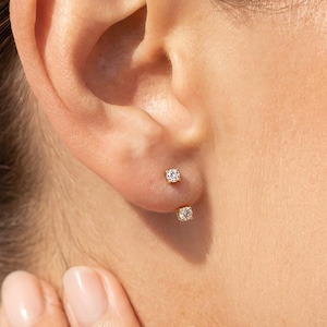 May include: Close-up of a person's ear with two small, round diamond stud earrings. The earrings are set in gold and are positioned on the earlobe. The skin tone is fair, and the background is blurred.
