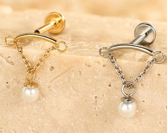 Könnte beinhalten: Zwei Ohrstecker, einer goldfarben und einer silberfarben, jeweils mit einer kleinen Kette und einer hängenden Kunstperle. Die Ohrringe werden auf einer strukturierten, hellbraunen Oberfläche präsentiert, die ihr zartes Design und die kontrastierenden Farben hervorhebt.