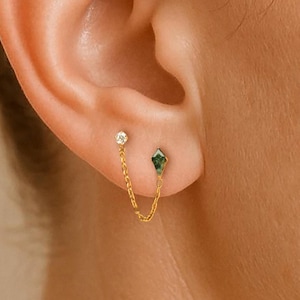 May include: A pair of gold earrings with a chain. One earring is a small, round, clear stone, and the other is a green, diamond-shaped stone. The earrings are connected by a delicate gold chain. The earrings are worn on an ear.