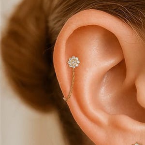 May include: Close-up of an ear with two gold-toned flower earrings. Each flower is set with small, sparkling stones. One earring has a chain that hangs from the upper ear, adding a touch of elegance to the design.