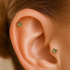 May include: Close-up of an ear with two small, green, four-leaf clover stud earrings. The earrings are set in a gold or silver metal. The ear is positioned on the right side of the frame, with the hair out of focus in the background.