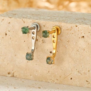 May include: A pair of earrings, one silver and one gold, each featuring a small green gemstone at the top and bottom. The earrings are displayed on a textured, light-colored block, with a blurred, neutral background.