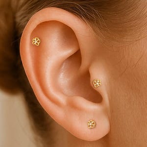 May include: Close-up of an ear adorned with three small, gold-colored stud earrings. Each earring features a delicate, floral design. The earrings are placed in the lobe and upper ear cartilage. The skin tone is fair, and the hair is pulled back.