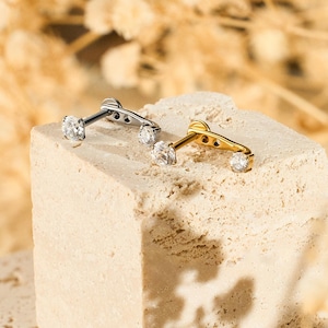 May include: Two pairs of earrings with a three-stone design. One pair is silver-toned, and the other is gold-toned. Each earring features three clear, round gemstones. The earrings are displayed on a textured, light-coloured block.